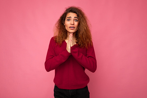 Photo of young emotional beautiful brunette curly woman with sincere emotions wearing trendy pink pullover isolated over pink background with free space and holding hands in pray gesture.