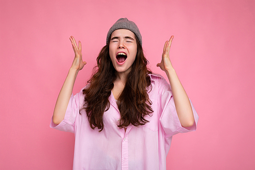 Beautiful emotional happy joyful young curly brunette woman wearing pink shirt and grey hat isolated on pink background with copy space and shouting.