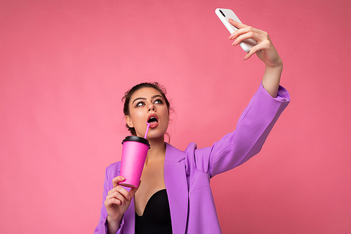 Close-up portrait of a beautiful young brunet woman in fashionable purple suit on a pink background in studio in a holding a popular phone and taking selfie photo and drinking through a straw beverage. Copy space