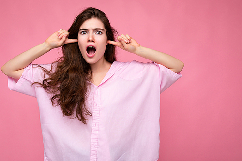 Portrait of young emotional winsome pretty curly brunet woman with sincere emotions wearing casual pink shirt isolated over pink background with copy space and covering ears with fingers and yelling.