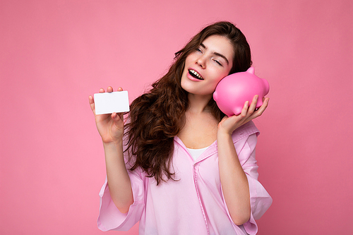 Attractive happy young brunette woman wearing shirt isolated on pink background with empty space and holding pink pig money box and credit card for mockup. Bank concept.