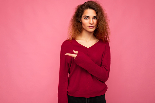 Photo of young beautiful brunette wavy woman with sincere emotions wearing casual pink sweater isolated over pink background with copy space and pointing at free space for mockup.