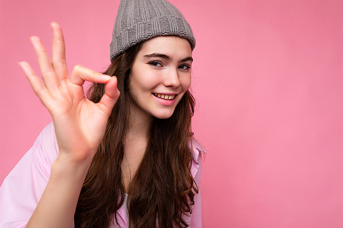 Closeup of adult positive happy winsome charming curly brunette woman with sincere emotions wearing stylish pink shirt and gray hat isolated on pink background with copy space and showing ok gesture. It's fine.