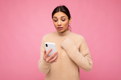 Attractive cute young shocked brunet woman wearing beige warm sweater standing isolated over pink background surfing on the internet via phone looking at mobile screen and keeping hands on heart.