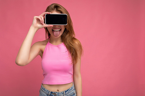 Photo of beautiful positive young blonde woman wearing pink top poising isolated on pink background with empty space holding in hand and showing mobile phone with empty display for mockup and showing tongue.