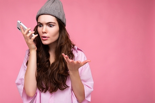 Emotional attractive young brunette woman wearing stylish pink shirt and grey hat isolated over pink background holding in hand and using mobile phone communicating and recording voice message explaining smth.
