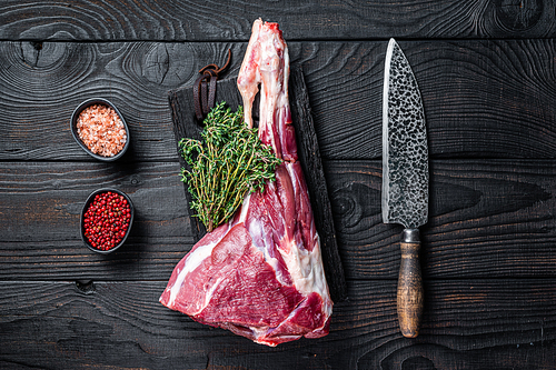 Uncooked Raw goat leg with herbs on butcher cutting board. Black wooden background. Top view. Copy space.