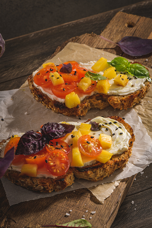 Italian bruschettas with roasted tomatoes, mozzarella cheese, pineapple slices and herbs on a wooden cutting board.