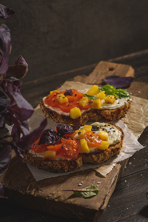 Italian bruschettas with roasted tomatoes, mozzarella cheese, pineapple slices and herbs on a wooden cutting board.