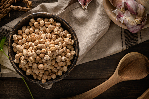 Raw chickpeas in a ceramic bowl, garlic and a wooden spoon on a dark background. View from above.