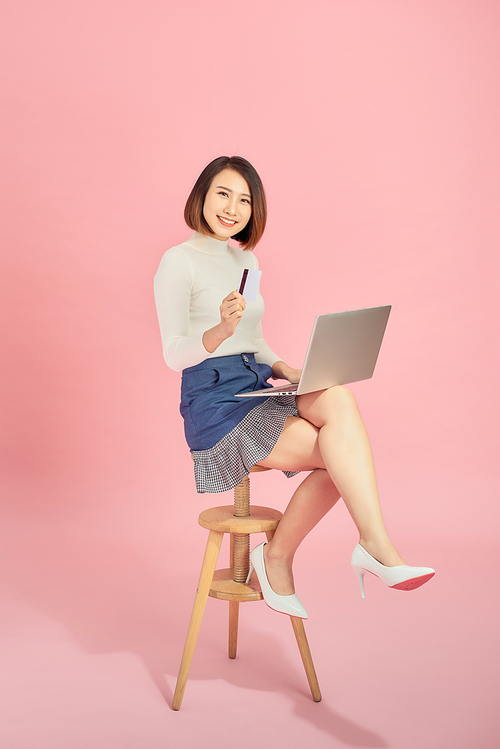 Young Asian woman using credit card to payment online with her laptop. Isolated on pink background.