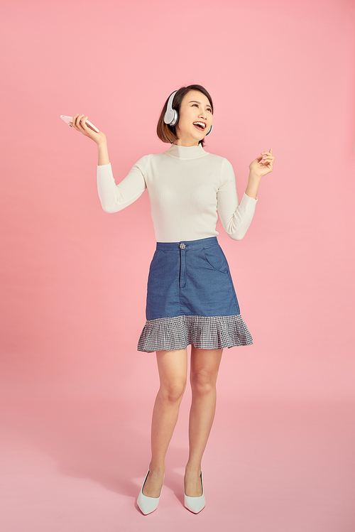 Full length portrait of a happy asian businesswoman listening to music with headphones while dancing isolated over pink background