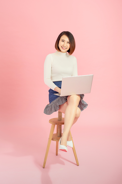 Young beautiful Asian businesswoman using laptop while sitting on chair. Isolated on pink background.