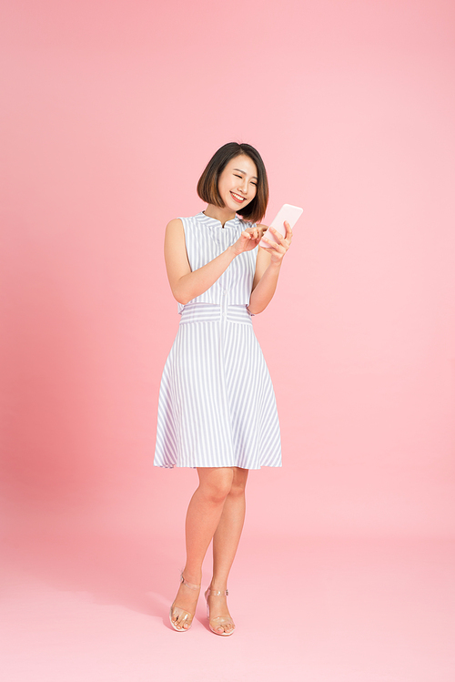 Full length portrait of a pretty young woman standing isolated over pink background, using mobile phone