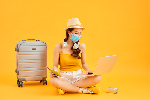 Young woman using laptop, wearing face mask while waiting for flight with her suitcase over orange background.