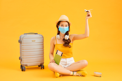 Young woman waiting for flight, sitting on floor near her suitcase, wearing face mask to prevent coronavirus.