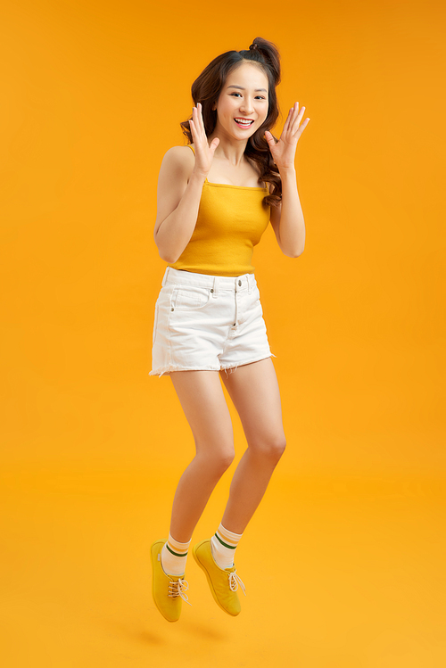 Full length portrait of a cheerful young asian woman celebrating success while jumping isolated over yellow background
