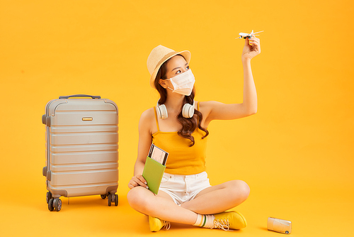 Young Asian woman sitting on the floor and holding passport, airplane while traveling and wearing face mask. Coronavirus concept.
