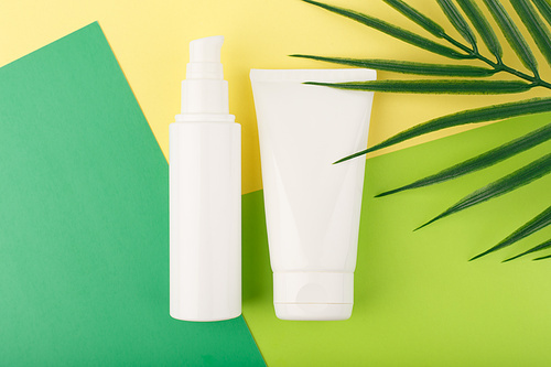 Cosmetic products in white unbranded tubes on green and yellow paper with palm leaf. Set of face cream, lotion or gel for skin cleaning, exfoliating, moisturizing and nourishing