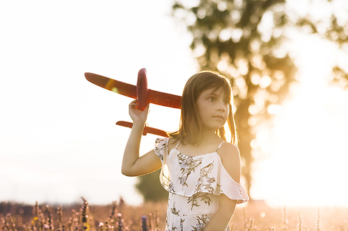 Portrait happy kid with toy airplane against orange sun summer sky background. Girl playing airplane on a summer lavender field. Best childhood concept.
