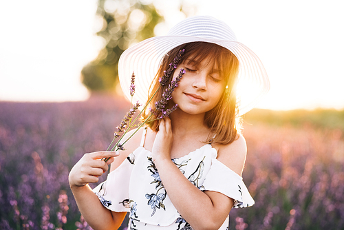 Portrait charming young girl in straw hat. Little girl with a bouquet of summer wildflowers. Childhood concept. Girl holding a bouquet of lavender. Kid girl in a white hat at the lavender field