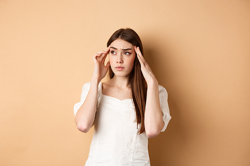 Troubled young woman feel sick, touching head temples, having headache, looking at upper left corner with tensed and distressed face, standing on beige background.