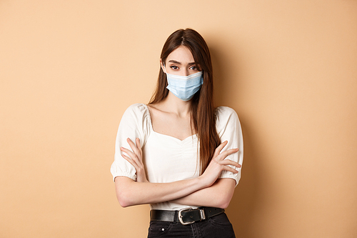 Covid-19 and lifestyle concept. Arrogant woman in medical mask frowning, cross arms on chest and looking confident at camera, beige background.
