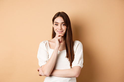 Smiling young european woman in dress, having an idea, listening with interest, touching chin and  intrigued, standing against beige background.