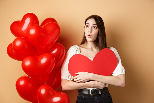 Valentines day and love concept. Intrigued tender girl hugging big red heart cutout and stand near balloons, looking aside at logo, saying wow, beige background.