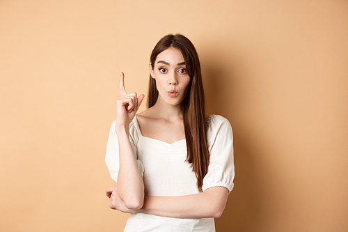 Creative girl pitching an idea, raising finger in eureka gesture, saying her plan, having a suggestion or solution, standing in dress on beige background.