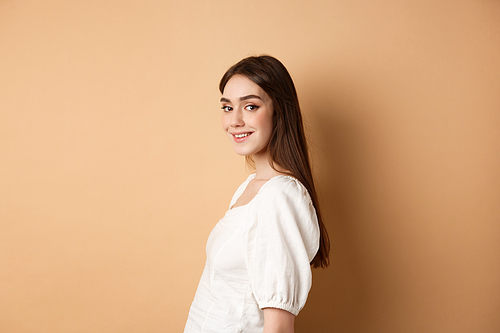 Profile of pretty european girl turn head at camera and smiling coquettish, standing in white blouse against beige background.