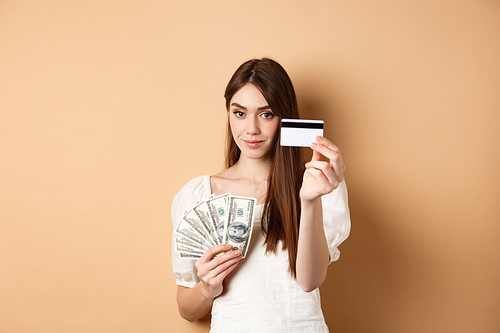 Young woman showing plastic credit card, prefer contactless payment instead of dollar bills, standing on beige background.