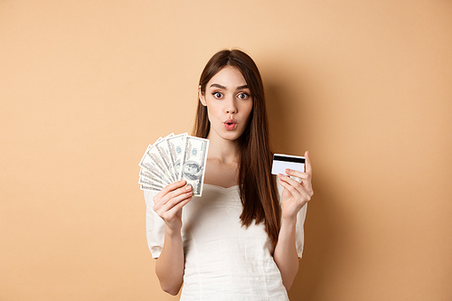 Excited girl showing dollar bills and plastic credit card, saying wow with amazed face, standing on beige background.
