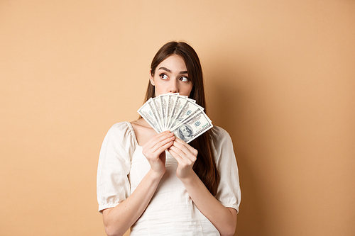 Thoughtful girl holding dollar bills and looking at empty space, thinking what to buy on money, plan shopping, standing on beige background.