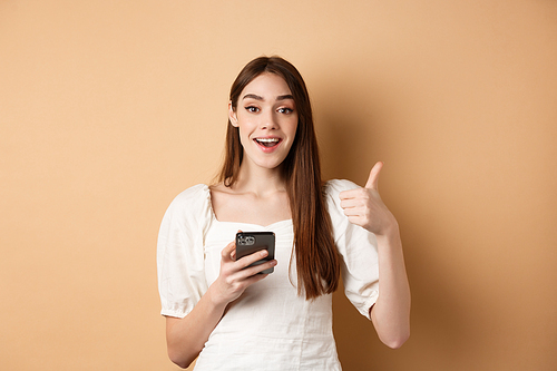 Online shopping concept. Happy young woman showing thumb up and using cellphone, smiling pleased, standing on beige background.