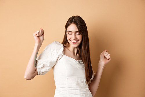 Carefree woman dancing and having fun, close eyes and smiling, standing on beige background.