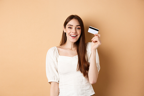 Cheerful young woman got her plastic credit card and smiling satisfied, standing pleased on beige background. Copy space