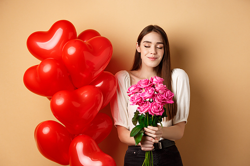 Romantic girlfriend smelling roses, receive flowers from lover on Valentines day, standing near red hearts balloons and smililng, beige background.