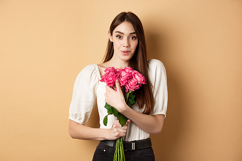 Valentines day. Surprised tender girl thanking for flowers and smiling, holding red roses and smiling grateful, standing on beige background.