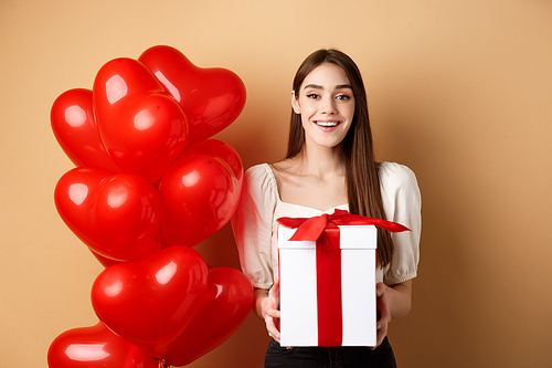 Romantic girl standing near heart balloons and holding surprise gift on Valentines day, smiling at camera happy, standing on beige background.