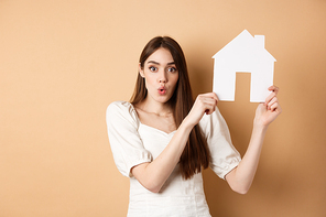 Real estate. Excited woman showing paper house cutout and saying wow with amazed face, buying property, standing on beige background.