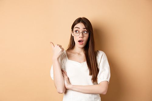 Excited girl in glasses pointing left, looking surprised at banner, standing on beige background.