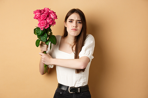 Happy valentines day. Angry girl hitting someone with flowers, swinging bouquet of roses at boyfriend, fighting with lover, standing on beige background.