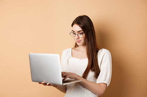 Young woman in glasses working on laptop, reading computer screen and smiling, standing on beige background.