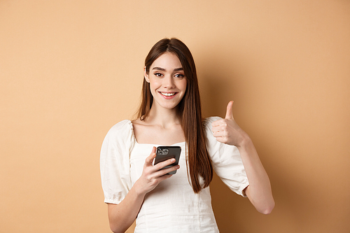 Online shopping concept. Smiling happy woman showing thumb up after using smartphone, holding phone and say yes, standing on beige background.