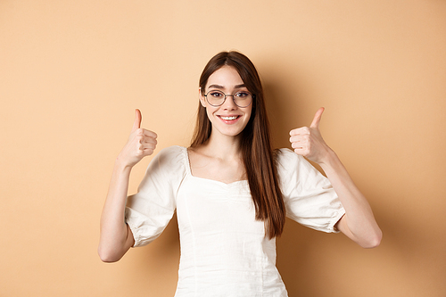 Happy girl in glasses show thumbs up, recommend eyewear store, smiling satisfied, standing on beige background.