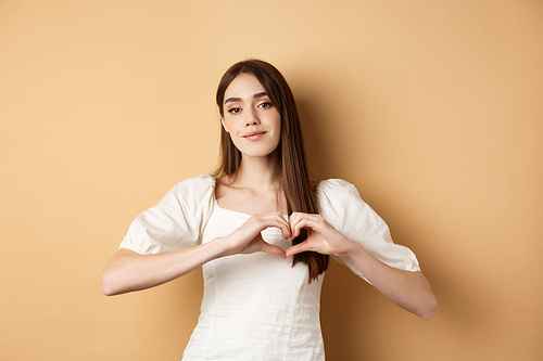 Valentines day and romance concept. Romantic caucasian girl in white dress showing heart gesture and smiling, say I love you, standing over beige background.