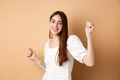 Happy woman dancing and having fun, close eyes and smiling, standing on beige background.