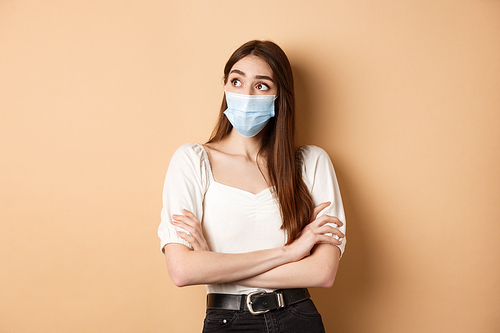 Pandemic and healthcare concept. Thoughtful girl in face mask looking at empty space logo, standing curious on beige background.