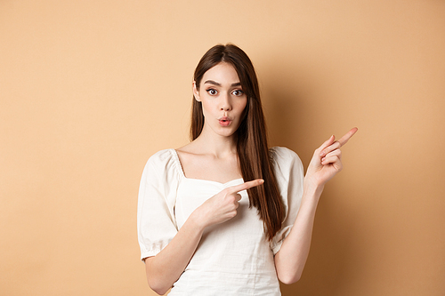 Wow look here. Impressed caucasian woman in white dress pointing fingers right and look curious, asking question about product, beige background.